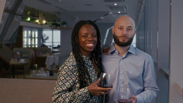 Medium copy space portrait of elegant bicultural couple with wine glasses looking at camera at restaurant with panoramic view