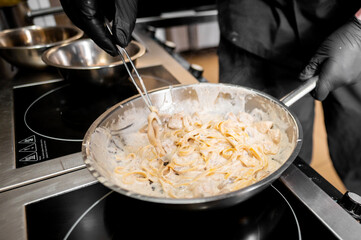  A chef in black gloves skillfully stirs creamy pasta in a stainless steel pan on a stovetop,...