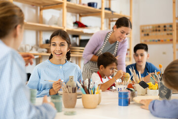 Joyful children, together with the teacher, paint ceramics with brushes at the table. Master class for children on pottery