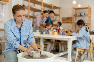 Concentrated teenage girl working at potter's wheel in taller workroom with other circle members