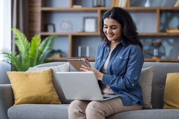 Young smiling Indian woman in casual clothes sitting on sofa, using laptop, making notes in notebook