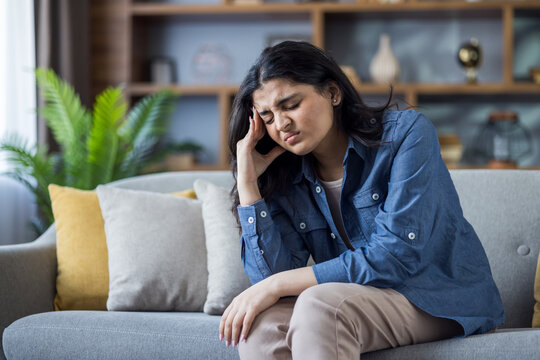 Suffering from headache and pressure, Indian young woman sits on sofa at home and grimaces holding her head