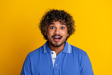 Cheerful young man with curly hair expressing surprise and happiness against a bright yellow...