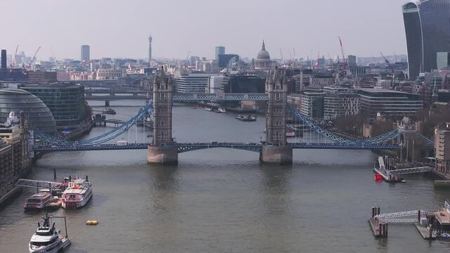 Aerial view of Tower Bridge with vehicles and pedestrians in motion, boats on the River Thames, and landmarks like St. Paul's Cathedral in the background.