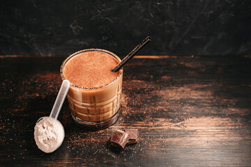 Healthy protein milkshake in a glass with a straw, a measuring scoop of whey protein powder, and chocolate pieces on a dark wooden background
