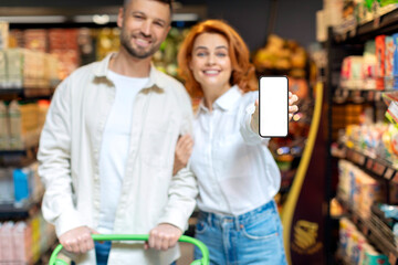 Grocery shopping app. Happy couple showing smartphone blank screen while shopping in supermarket, selective focus on phone, mockup