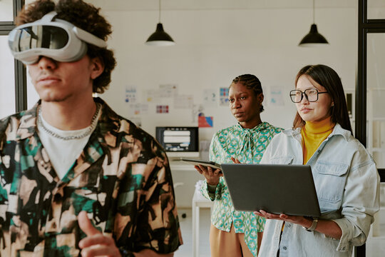 Medium shot of concentrated man playing VR game while two biracial female gamedevs holding devices and watching him playing