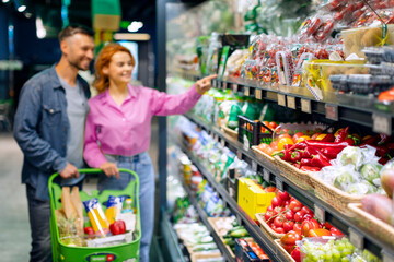 Focus on fresh vegetables on shelf in grocery store department, couple choosing groceries in supermarket on background