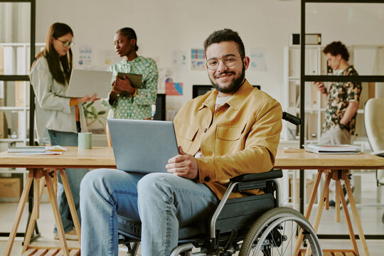Portrait of handsome young IT specialist with disability holding laptop of knees smiling at camera while his colleagues working in background