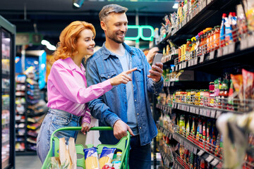 Couple using smartphone while buying food in supermarket, checking their list and choosing products, standing with shop cart in groceries store