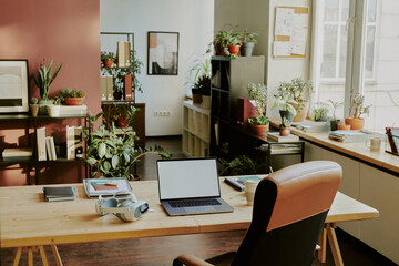 High angle shot of cozy it office equipped with work table covered with copybooks and digital devices, spacious room decorated with green plants in pots