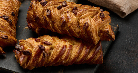 Three golden brown maple pecan braid pastries are arranged on a dark, textured surface. Braided dough is flaky and topped with pecans