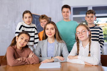 Group photo of teacher and preteen schoolchildren sitting at table and some staying behind in classroom