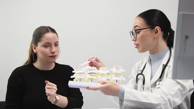Female doctor explains human spine model to young patient in medical office. Concept of back treatment, diagnostics and physiotherapist consultation.