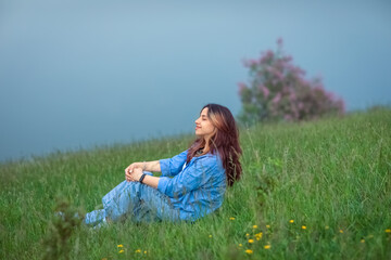 Fototapeta premium a young woman sitting on a lush green grassy hill near a water. Her eyes are closed, and she has a peaceful, content expression on her face. Her arms are wrapped around her knees in a relaxed pose