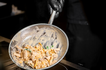 A chef in black gloves tosses creamy mushroom fettuccine pasta in a metallic pan, showcasing an appetizing Italian dish in a professional kitchen setting.