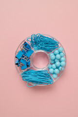 A creative arrangement of blue office supplies, including paper clips, rubber bands, and push pins, neatly organized in a circular container against a soft pink background. A colorful and stylish