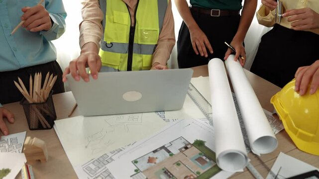 Architect and safety engineer discussion about building blueprint displaying on laptop on table with architectural document and wooden block scatter around at modern office. Closeup. Delineation.