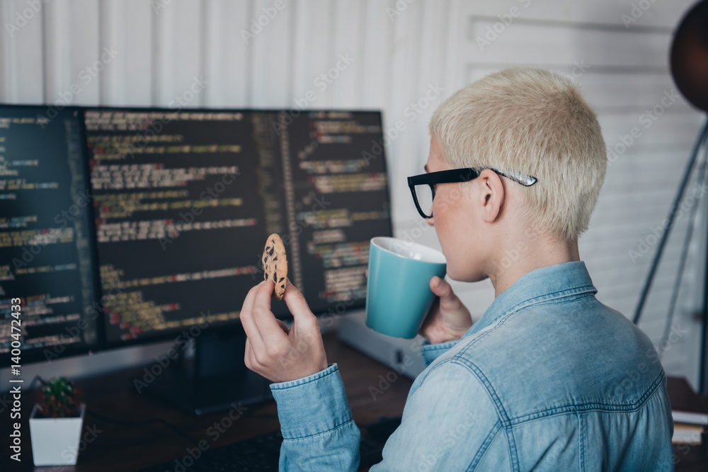 Poster blond woman in denim shirt coding at computer while enjoying coffee and cookie