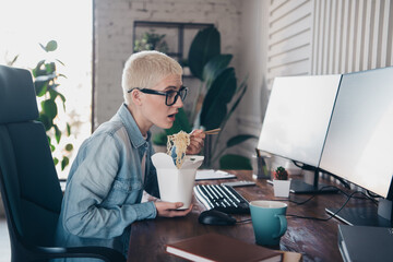 Young woman in casual denim shirt eating noodles while working with computers at home office