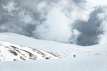 Hiker with backpack and trekking poles walking across snow-covered slopes in Sierra de Gredos, Spain, under dramatic storm clouds in a remote and rugged alpine winter setting