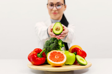 A nutritionist in a lab coat with an avocado in hand sits at a table of fresh fruits and vegetables...