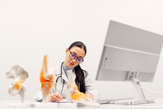 A female physical therapist sits at a table at a computer next to anatomical training models of joints for demonstration. Health Concept.
