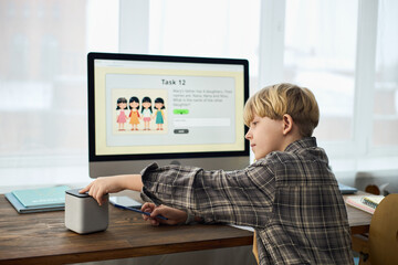 Young boy engaged in online learning activity, interacting with tablet, smart home device on table. Computer screen displaying visual learning task