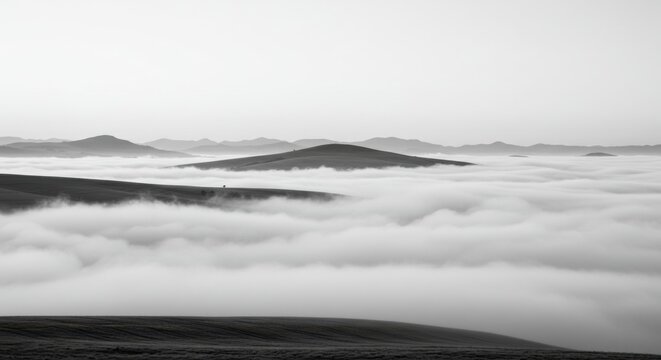 A monochrome landscape photograph captures rolling hills emerging from a dense layer of fog or low clouds, with distant mountain peaks visible on the horizon under a soft, overcast sky