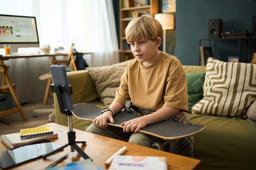 Young boy sitting on couch with skateboard on lap, engaging with a smartphone on a tripod in brightly lit living room showcasing relaxed atmosphere © DragonImages