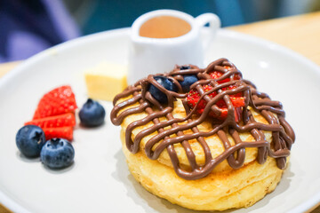A waffle topped with chocolate drizzle, strawberries, and blueberries, served with syrup, butter, and extra fruits on a plate.