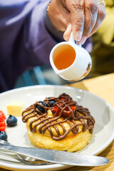 A close-up of a person pouring syrup onto a plate of gourmet pancakes topped with chocolate drizzle and fresh fruit, such as strawberries and blueberries.