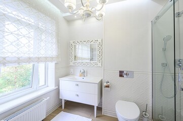 Bright, white bathroom with classic details. Features a vanity, ornate mirror, toilet, and a glass-enclosed shower. Natural light through a window adds to the airy feel.