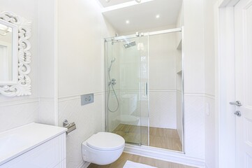 Bright, white bathroom with classic details. Features a vanity, ornate mirror, toilet, and a glass-enclosed shower. Natural light through a window adds to the airy feel.