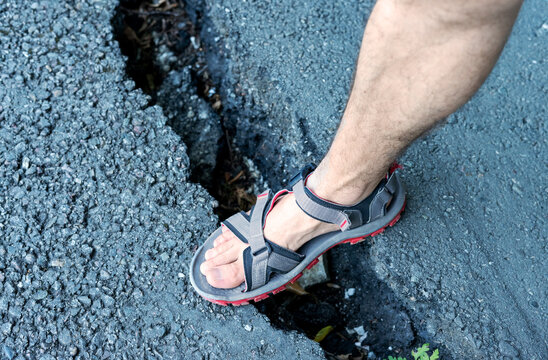 Foot in grey sandal steps into crack on an asphalt road, infrastructure damage, posing potential hazards for pedestrians Outdoor setting with visible gravel