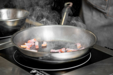 Close-up of a stainless steel pan on an induction cooktop with sizzling bacon slices, steam rising,...