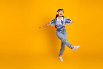 Cheerful young woman wearing striped shirt and headphones dancing playfully on yellow backdrop...