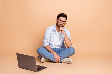 Young man with beard sitting casual fashionably against beige background, wearing striped shirt and glasses, thinking expression
