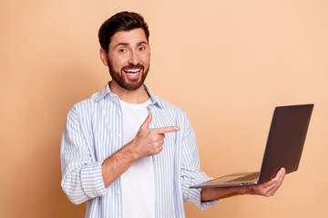 Cheerful young man in casual outfit holding a laptop and making a gesture against a beige wall