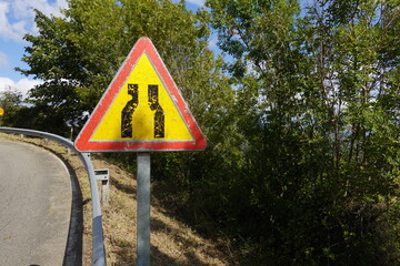 A worn warning sign showing road narrowing stands beside a rural road, surrounded by greenery and trees, with faded paint from exposure.