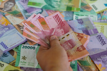 Hands holding several hundred thousand rupiah bills with a background of various types of Indonesian money. Suitable for background, finance, banking, cash, savings.