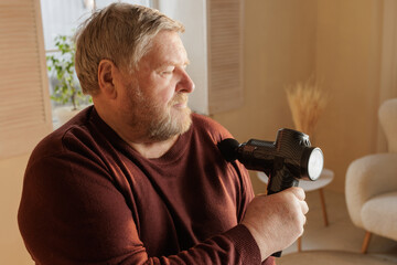 Senior man applying massage gun on shoulder for pain relief at home.