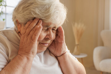 Senior woman holding head in pain, struggling with headache or stress indoors.