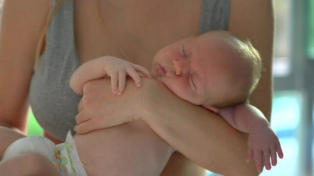 A young mother gently rocks her newborn baby in her arms by a sunlit window on a warm summer day as the baby slowly wakes up. A tender moment of love, bonding, and natural warmth in the glow of