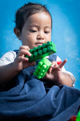 Joyful toddler playing with building blocks while sitting against a blue background. The child's expression shows curiosity and delight in this candid indoor moment.