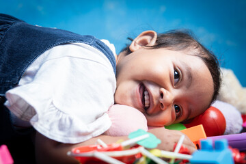 An Asian toddler girl lies on her stomach hugging a teddy bear, surrounded by toys. She has a soft smile with a hint of tiredness, capturing a quiet moment after playtime.