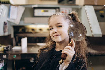 A young girl with long hair holds a magnifying glass, smiling confidently. She is in a kitchen...
