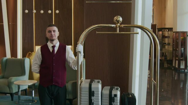 Portrait of bellboy in elegant uniform standing in hotel lobby next to trolley with luggage, looking at camera with confidence
