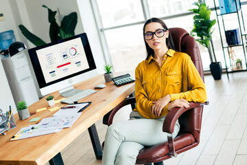 Young businesswoman in a modern office setting, seated confidently at her desk surrounded by...