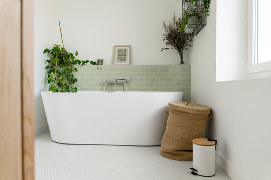 Salle de bain r&eacute;nov&eacute;e avec baignoire &icirc;lot blanche, carrelage en zellige vert clair, plantes vertes, panier &agrave; linge en osier et meuble en bois.